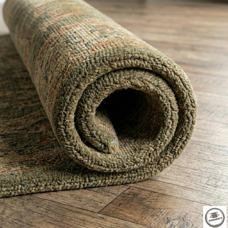May include: Rolled-up olive green area rug with a textured surface. The rug has a pattern of brown and orange accents. The rug is on a wooden floor.