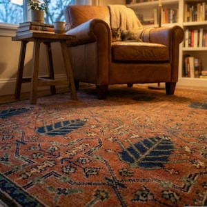 Peut inclure: Tapis de style ancien avec un motif détaillé dans des tons rouille, bleu marine et vert. Le tapis présente un motif central avec des motifs en forme de feuilles et des bordures complexes. Un fauteuil en cuir et une table d'appoint en bois sont en arrière-plan.