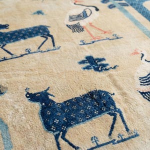 May include: Beige rug featuring a repeating pattern of blue deer and birds. The rug has a detailed design with floral and tree motifs. The deer are dark blue with white dot patterns. The birds are white with blue and pink accents.