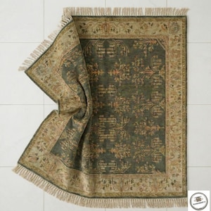 May include: An area rug with a dark green central field and a beige border. The rug features an intricate floral pattern in shades of beige and gold. The edges have a fringe detail. The rug is partially folded, revealing its texture.