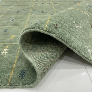 May include: A sage green area rug with a geometric pattern and rolled edge. The rug features a grid design with yellow lines and small, stylized tree and bird motifs in white, brown, and blue. The rug is made of a textured material.