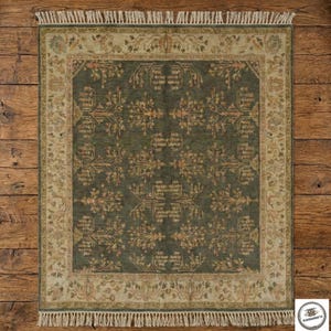 May include: A square, dark green rug with an intricate floral pattern in shades of tan and gold. The rug has a beige border with a repeating design and fringe along the edges. The rug is displayed on a wooden floor.