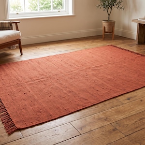 May include: A rectangular area rug in a solid burnt orange color featuring a textured surface with small raised tufts throughout. The rug has short, straight fringe detailing along the two shorter ends and lies flat on a wooden floor surface.