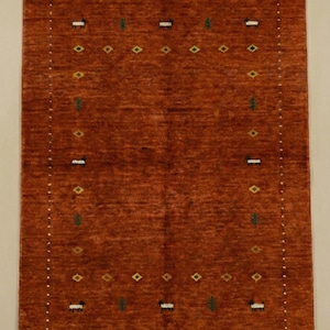 May include: A rectangular, rust-colored rug with a patterned border. The border features small white dots, with alternating diamond and tree-like designs in green, yellow, and white. The rug's central field is a solid, textured rust color.