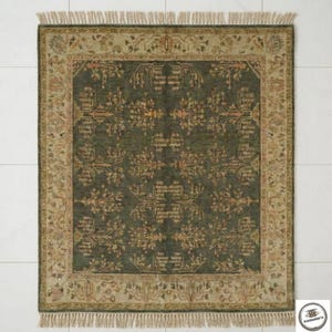 May include: A rectangular, dark green rug with a detailed floral pattern and a beige border. The rug has a fringe on the edges and is placed on a white tiled floor. The rug's design features symmetrical floral motifs.