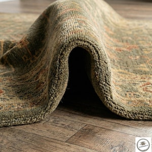 May include: Close-up of a rolled-up, olive green area rug with a distressed, patterned design. The rug's texture is visible, with a mix of olive, brown, and rust-colored details. The rug is on a wooden floor.