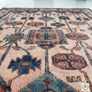 May include: A close-up of a vintage rug with a detailed geometric pattern. The rug has a soft pink background with intricate designs in shades of blue, orange, and brown. The design includes floral and geometric shapes.