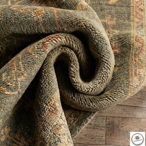May include: Close-up of a rolled-up, olive green rug with a detailed, textured pattern. The rug features a border with a repeating design in shades of yellow, red, and beige. The rug's surface has a soft, plush appearance, suggesting a comfortable texture.
