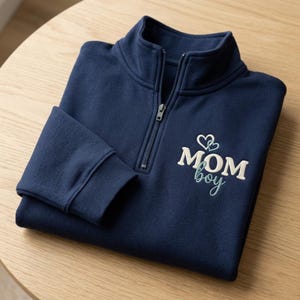 May include: A navy blue quarter-zip pullover featuring embroidered text that reads MOM in cream block letters and boy in teal cursive script, accented by two small teal heart outlines positioned above the word MOM on the left chest area.