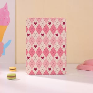 May include: A rectangular e-reader case featuring a repeating argyle pattern in shades of light pink and cream. The design is accented with small, dark pink hearts positioned at the intersections of the thin, dark brown diamond-shaped grid lines.