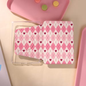 May include: A rectangular case featuring a pink and white argyle pattern with small dark pink hearts centred within each diamond shape. The case has a clear plastic base and a hinged lid decorated with the repeating heart and diamond design.