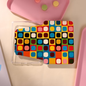 Può includere: Una custodia rettangolare caratterizzata da un vivace motivo a griglia di quadrati multicolori con angoli arrotondati nelle tonalità del blu, giallo, rosso, rosa, bianco e marrone scuro. La custodia è parzialmente aperta, rivelando una struttura interna in plastica trasparente.