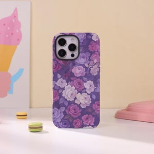 May include: A rectangular smartphone case featuring a repeating floral pattern of roses in various shades of purple, lavender, and white against a dark purple background, with a cutout for a triple-lens camera system on the upper left corner.