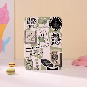 May include: A sage green e-reader cover decorated with various black and white stickers, including a ghost reading a book, a soda can labelled Plot Twist Pop, a Queen of Books playing card, and text reading In My Kindle Era and Just One More Page.