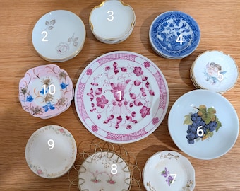 May include: A collection of ten small ceramic plates and saucers featuring various designs, including floral patterns, blue and white willow motifs, cherubs, and a central plate with a pink botanical print and a gold wire display stand.