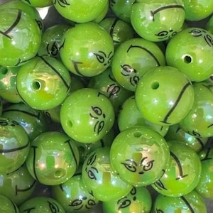 Pode incluir: Uma coleção de contas redondas verde-limão, cada uma com um gráfico preto impresso de uma expressão facial travessa com olhos estreitos e um sorriso. As contas têm um acabamento liso e brilhante e um orifício central para enfiar.