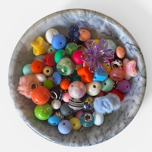 Può includere: Una ciotola di marmo piena di una varietà di perline e ciondoli colorati. L'assortimento comprende perline rotonde, a forma di stella e di fiore nelle tonalità dell'arancione, del blu, del rosa e del viola. Alcune perline hanno una consistenza pelosa e altre sono a righe.