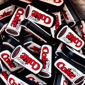 May include: A collection of small, rectangular shoe charms featuring the Diet Coke logo in red and white lettering on a black base. Each charm displays the brand name clearly, with a white background, red text, and a black border, stacked together.