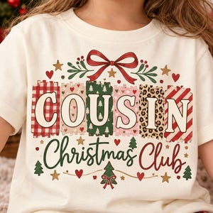 May include: A cream-colored short-sleeve t-shirt featuring a graphic with the text "COUSIN Christmas Club". The design includes a red bow, various patterned rectangular blocks behind the letters, small stars, hearts, and Christmas trees.