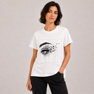 May include: A white short-sleeved t-shirt featuring a central rectangular graphic print. The design displays a detailed black ink illustration of an eye with eyelashes transitioning into bare tree branches and a flock of small birds flying away.
