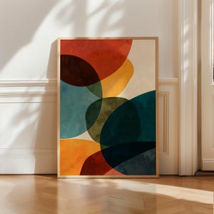 Op de afbeelding: Een rechthoekige wandkunstprint in een lichthouten lijst met een abstracte compositie van overlappende organische vormen in tinten terracotta, mosterdgeel, teal, donkerblauw en crème, wat een gelaagd aquareleffect creëert.