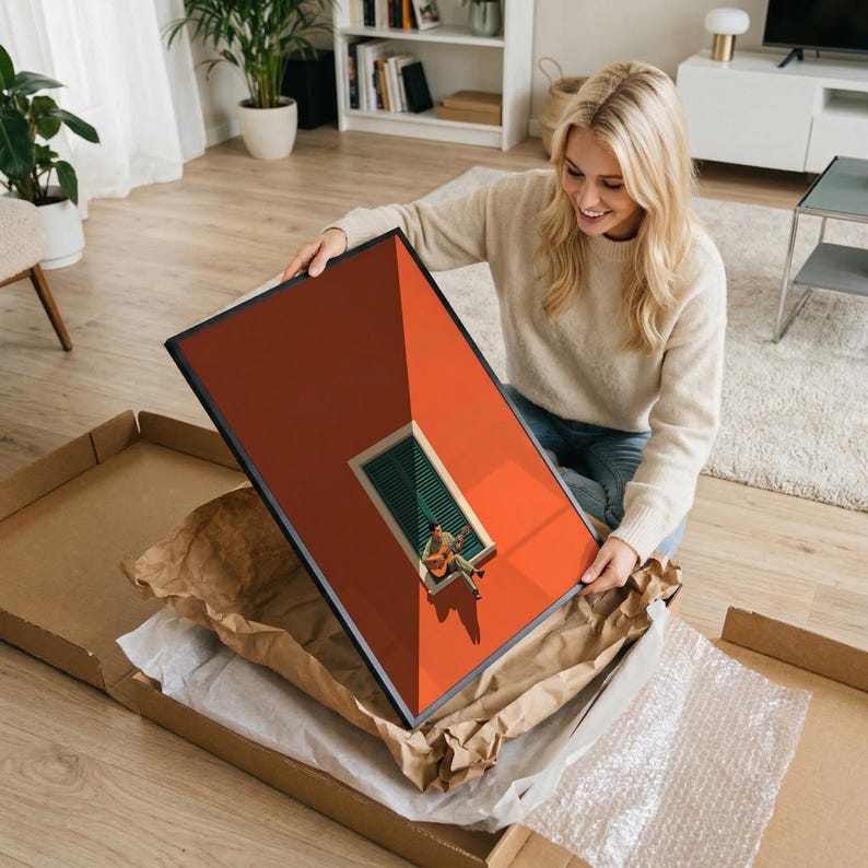May include: A rectangular framed wall art print featuring a vibrant orange wall with a central window that has green shutters. A person is sitting on the window ledge playing an acoustic guitar, casting a long shadow on the orange surface.