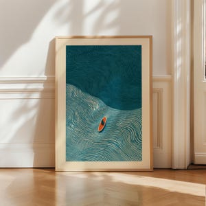 May include: A framed art print featuring an abstract illustration of a person on a surfboard in the ocean. The artwork uses shades of teal and cream to depict the water and a bright orange surfboard. The frame is a light wood color.