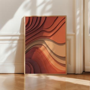May include: A framed abstract art print with layered, wavy forms in orange, tan, and brown hues. The artwork is displayed in a room with a wooden floor and white walls, with natural light creating shadows.