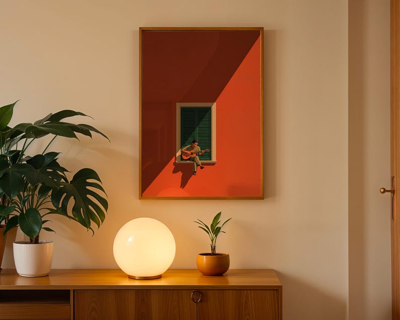 May include: A framed wall art print featuring a vibrant orange building facade with a central window. A person sits on the window ledge playing an acoustic guitar. The window has dark green shutters and is cast in a sharp, diagonal shadow.