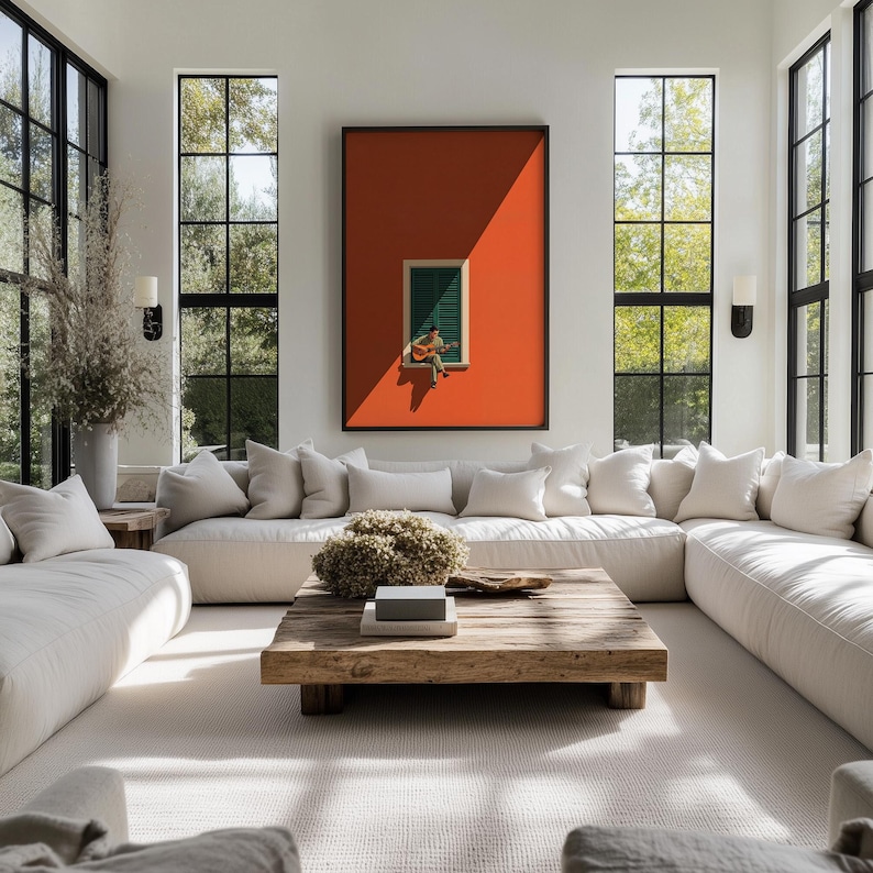 May include: A large rectangular framed wall art piece featuring a vibrant orange background with a central green shuttered window where a figure sits playing an acoustic guitar, displayed above a cream-colored sectional sofa and wooden coffee table.