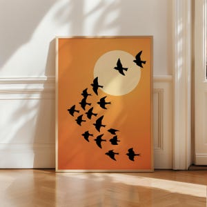 Könnte beinhalten: Ein rechteckiger Wandkunstdruck in einem hellen Holzrahmen mit einem orangefarbenen Farbverlaufshintergrund, der in eine blassgelbe, kreisförmige Sonne übergeht. Eine Schar schwarzer Vogelsilhouetten fliegt in einer diagonalen Formation.