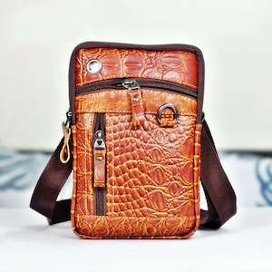 May include: A brown leather crossbody bag with a crocodile skin texture. The bag has multiple zippered compartments, a brown adjustable strap, and silver-toned hardware. The bag is designed for carrying essentials.