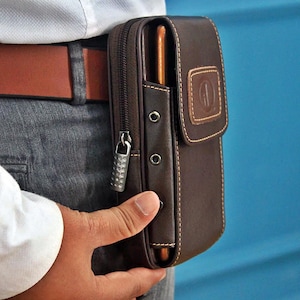 Puede incluir: Una funda de teléfono de cuero marrón con cierre de cremallera y trabilla para cinturón. La funda tiene una solapa con un logotipo cosido y costuras marrones claras en contraste. El teléfono es parcialmente visible en el bolsillo abierto.