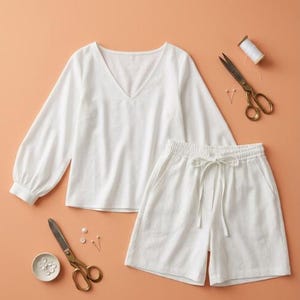 May include: A white long-sleeve top with a V-neckline and matching white shorts with an elastic waistband and drawstring. The set is accompanied by two pairs of metal scissors, a spool of white thread, several sewing pins, and small white buttons.