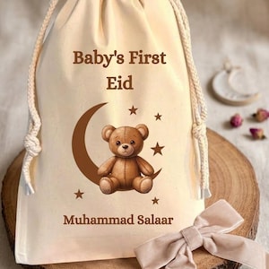 May include: A cream drawstring gift bag printed with "Baby's First Eid" and the name "Muhammad Salaar". The design includes a brown teddy bear on a crescent moon with stars. A beige bow sits on a wooden surface.
