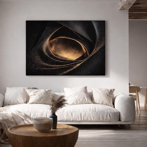 May include: Abstract canvas art featuring a swirling, dark composition with a golden center. The artwork, mounted on a wall, is the focal point of a living room with a white sofa, pillows, and a wooden coffee table. The room has a neutral color palette.