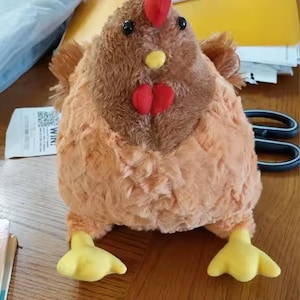 Puede incluir: Un juguete de peluche con forma de pollo con cuerpo marrón, pecho naranja y patas amarillas. Tiene cresta y barbilla rojas, y ojos negros. El juguete está sobre una superficie de madera.
