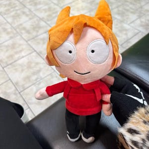 May include: A plush doll of a cartoon character with orange hair and large white eyes. The doll is dressed in a red hoodie, black trousers, and white trainers. The doll is approximately 20 cm tall.