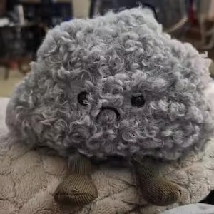 May include: A grey, cloud-shaped plush toy with a sad expression and small, stitched feet. The toy has a curly, textured surface and black button eyes. It is resting on a soft, light-coloured surface.