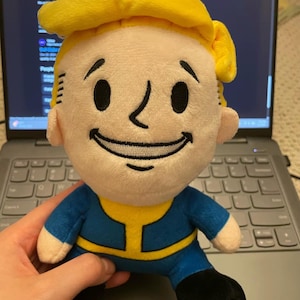 Könnte beinhalten: Plüschpuppe einer Figur mit gelber Kappe, blauem Overall und schwarzen Schuhen. Die Puppe hat ein lächelndes Gesicht mit großen schwarzen Augen und einer kleinen Nase. Die Figur stammt aus der Fallout-Videospielserie.
