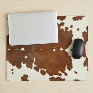 May include: A rectangular mousepad with a brown and white cowhide pattern. A silver laptop and a black wireless mouse are on the mousepad. The mousepad is on a light wood surface.