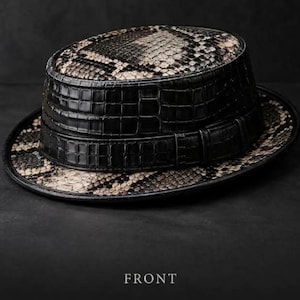 Op de afbeelding: Een fedora hoed met een kroon en rand in een beige en zwart slangenleerpatroon, gecontrasteerd door een brede, zwarte, krokodil-geëmbosseerde leren band rond de basis. Het woord FRONT staat in witte hoofdletters onder de hoed gedrukt.