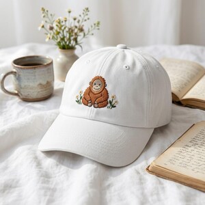 May include: White baseball cap featuring an embroidered orangutan design. The orangutan is brown with a smiling face, surrounded by embroidered flowers. The cap has a curved brim and a button on top.