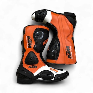 Könnte beinhalten: Paar orange-schwarze Motorradstiefel mit weißen Akzenten. Die Stiefel zeigen das KTM Racing Team Logo und Schutzplatten. Diese hoch geschnittenen Stiefel sind für das Motorradfahren konzipiert.