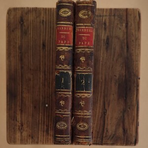 Op de afbeelding: Twee antieke boeken met versleten, bruine leren covers. De ruggen tonen de titel "BARRUEL DU PAPE" in rode en gouden letters. De boeken zijn gemarkeerd met "1" en "2" en liggen op een houten oppervlak.