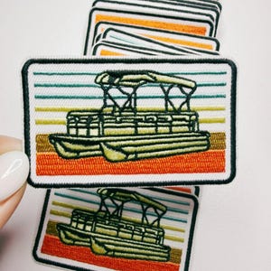 May include: Embroidered patch featuring a pontoon boat design. The patch has a rectangular shape with a black border. The boat is olive green, set against a backdrop of horizontal stripes in orange, brown, green, and white. A stack of similar patches is visible.