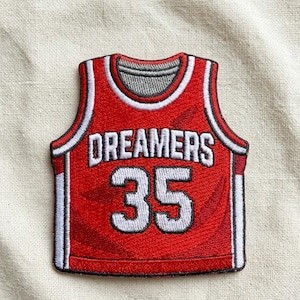 May include: Red jersey patch with the word "DREAMERS" in white block letters above the number "35". The jersey has white and black trim and a gray neckline. The patch is on a beige fabric background.