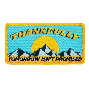 May include: Embroidered patch with a blue sky background, a yellow sun, and mountain silhouettes. The patch reads "THANKFULLY TOMORROW ISN'T PROMISED" in yellow text, framed by a gold border. A decorative item.