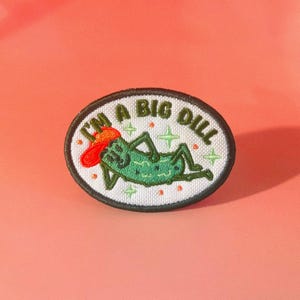 May include: Embroidered patch with a green pickle wearing a red cowboy hat, reclining on a white background. The patch reads "I'M A BIG DILL" in a playful font, surrounded by a black border. Green star and orange dot accents.