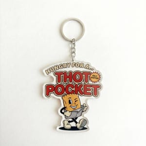 May include: A clear acrylic keychain with a cartoon image of a walking bread pocket reading a newspaper. The keychain has the words "HUNGRY FOR A... THOT POCKET" in red and white lettering. The keychain is attached to a silver metal keyring.